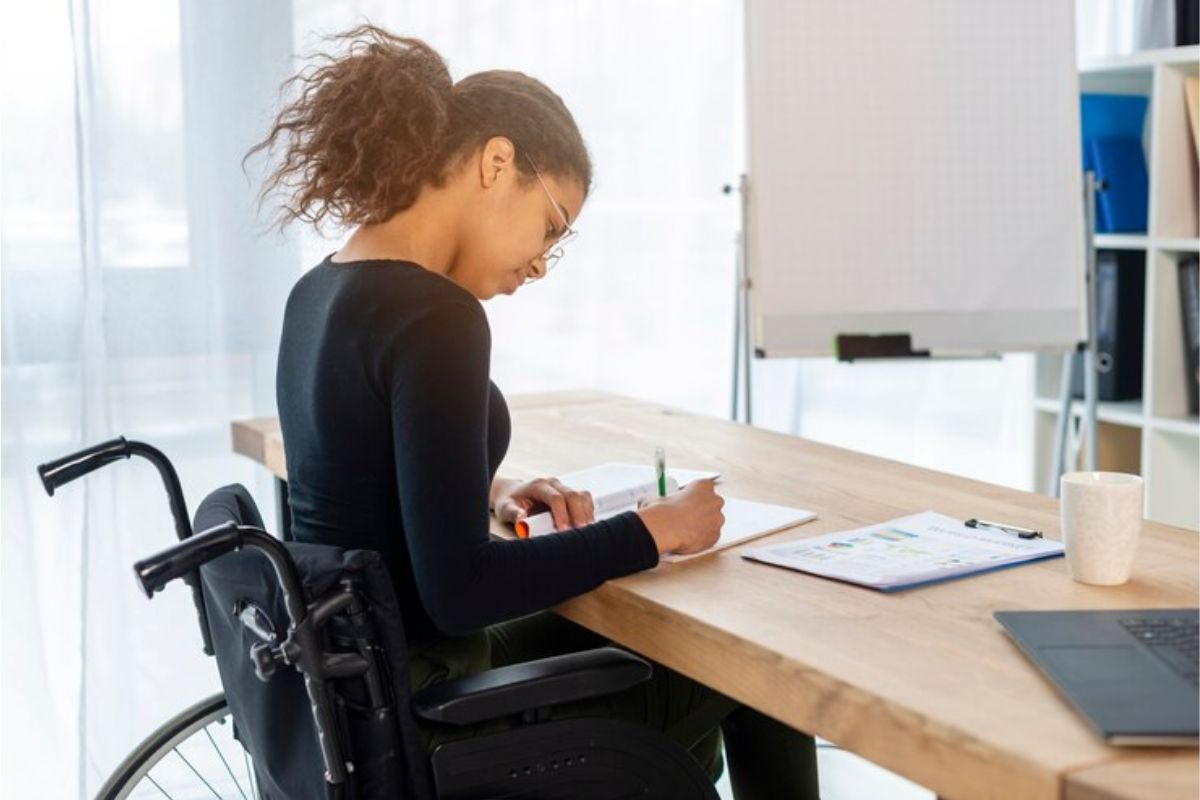 Uma mulher na cadeira de rodas trabalha em um escritório. Ela está escrevendo em seu caderno que está em cima da mesa