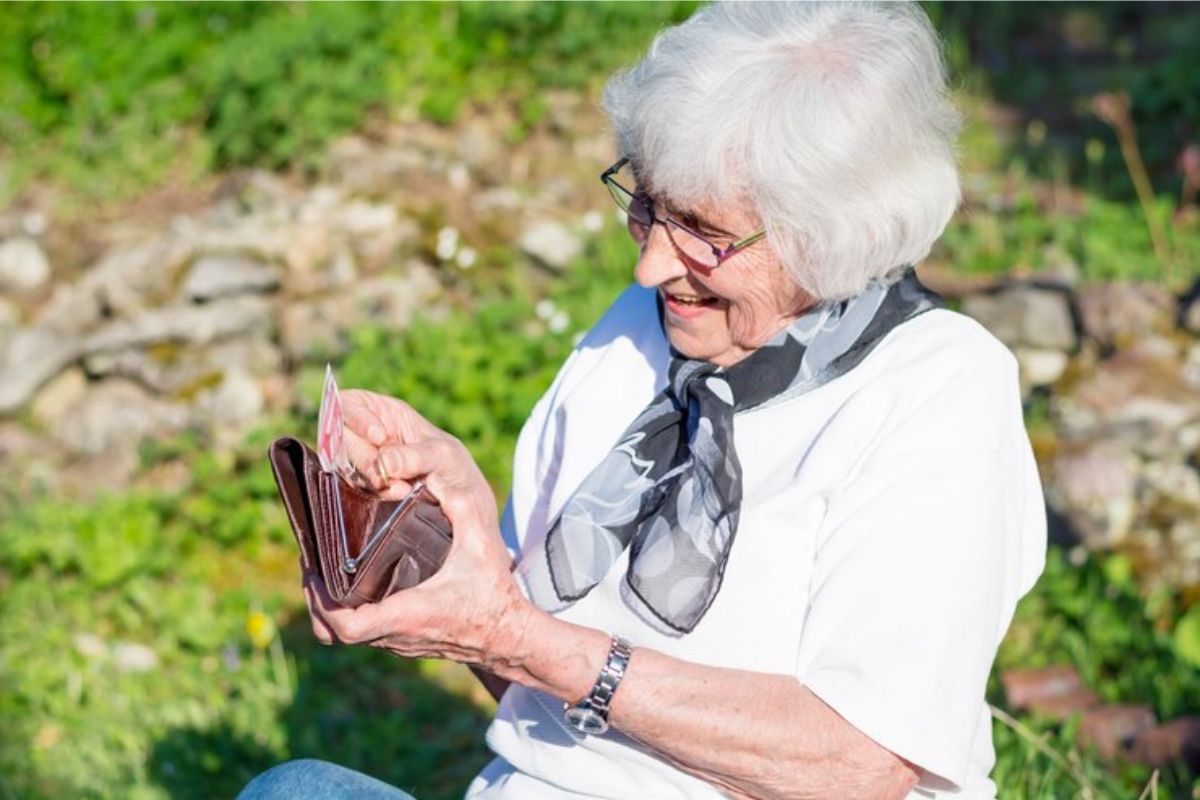 Uma senhora com a carteira na mão tirando dinheiro de dentro. Ela está sentada ao sol e feliz após ver a notícia de renda extra para quem mais precisa