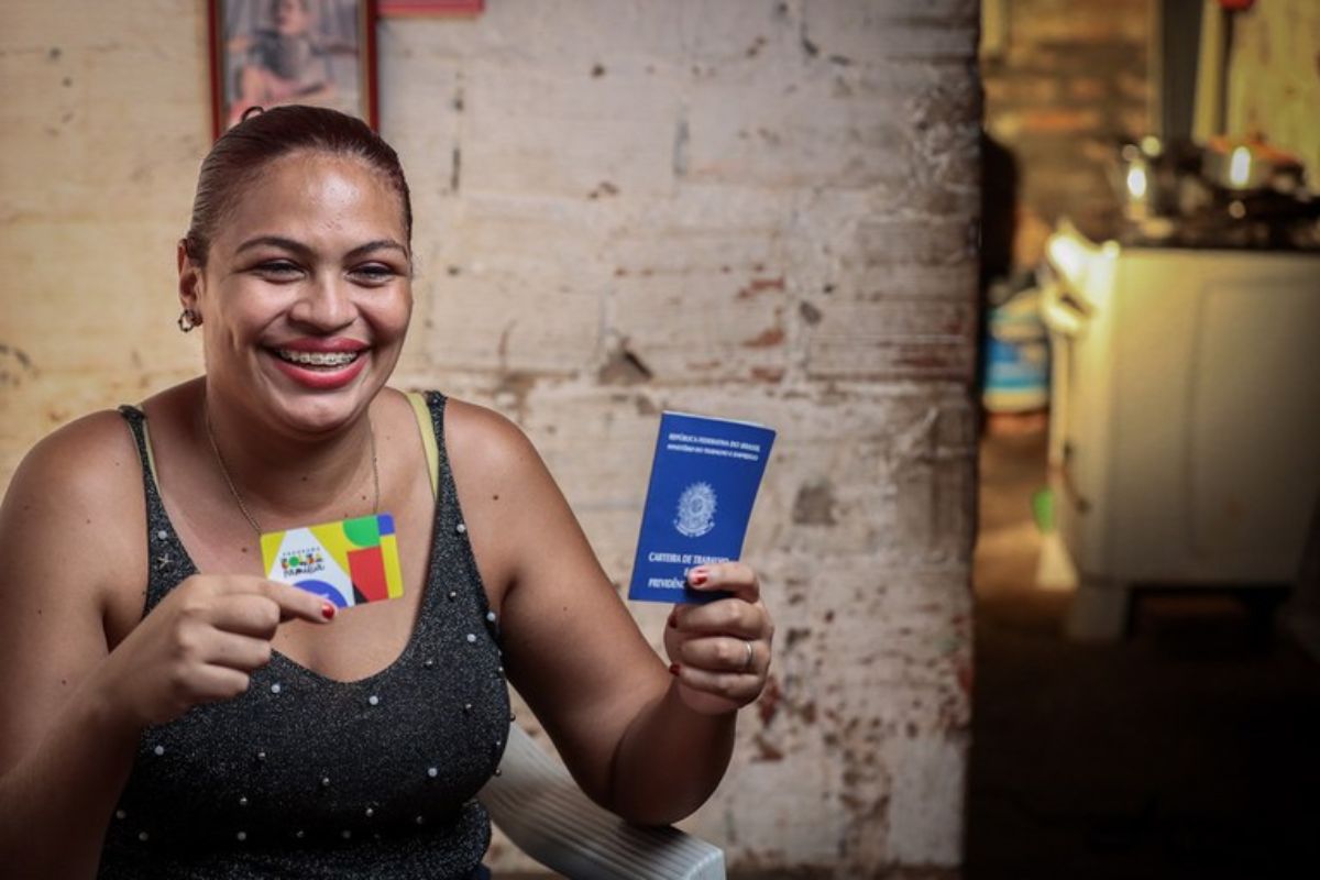 Uma mulher sorrindo com a carteira de trabalho na mão e o cartão do Bolsa Família. Ela comemora, pois, cmeça a festa do Bolsa Família