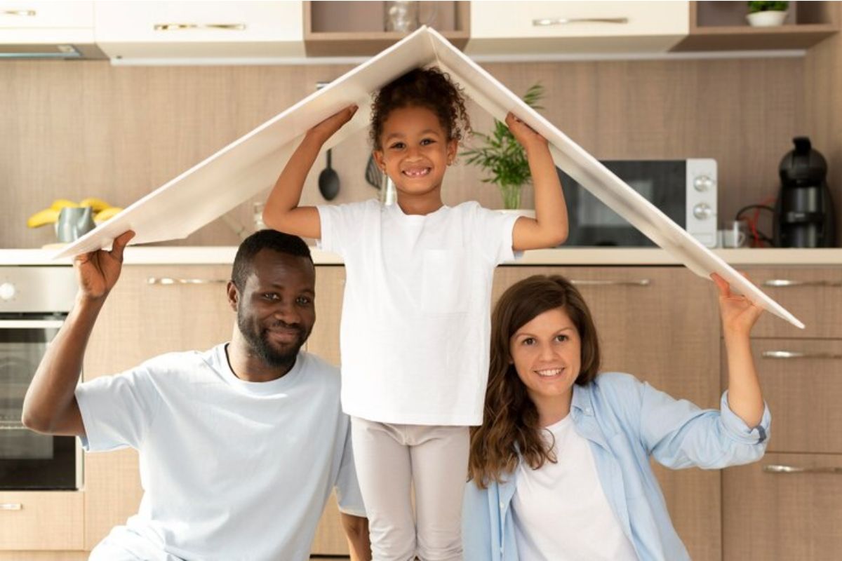 Uma família feliz em sua cozinha segura um papelão simulando um teto. Eles estão sorrindo após ver que o Governo Federal emite comunicado sobre novo plano habitacional 