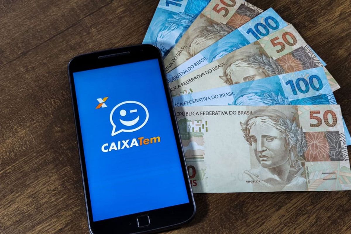 Imagem de um celular em cima de notas de R$ 50 e R$100. Caixa tem benefício de R$ 800