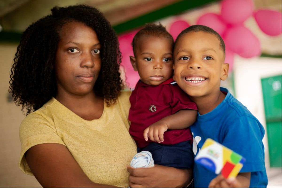 Uma mãe com um filho no colo e outro ao lado. Ela está com a cara emburrada, se questionando se o Bolsa Família bloqueado acumula ou é cancelado