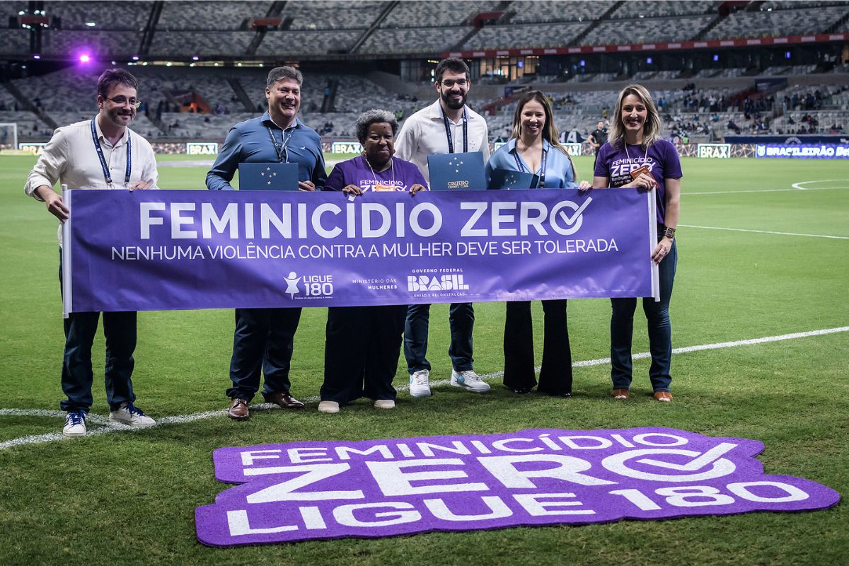 Três mulheres e três homens seguram uma faixa no campo de futebol referente à Campanha FEMINICÍDIO ZERO 