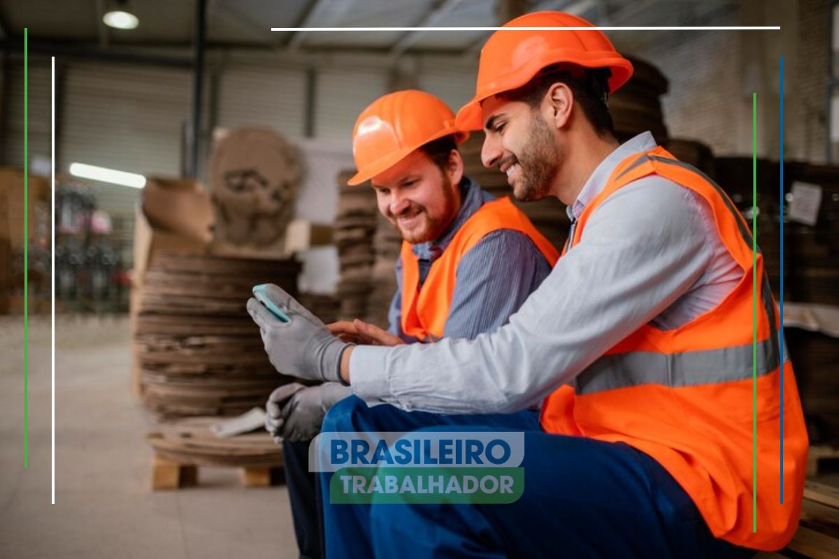 Dois colegas de trabalho com uniforme e capacete laranja observam em um smartphone o anúncio do Novo SALÁRIO MÍNIMO de R$ 1.509 aprovado