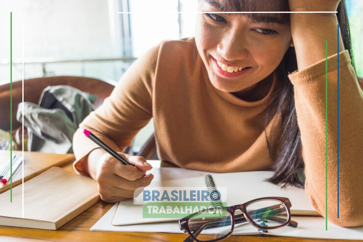 Uma aluna estudando em casa. Ela escreve em seu caderno e sorri depois de ver que São Paulo oferece Bolsa Trabalho com cursos gratuitos e auxílio 