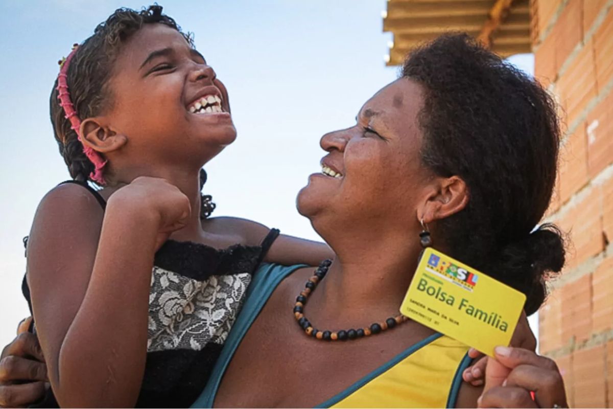 Uma mãe segura o cartão do benefício e sua filha no colo feliz ao ver que o Bolsa Família oferece descontos em imóveis