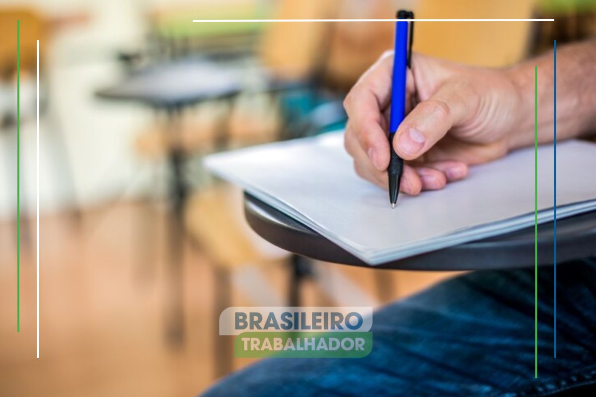 O braço de uma pessoa na mesa aparece escrevendo em uma prova. Ele participa do NOVO concurso em Conceição do Jacuípe