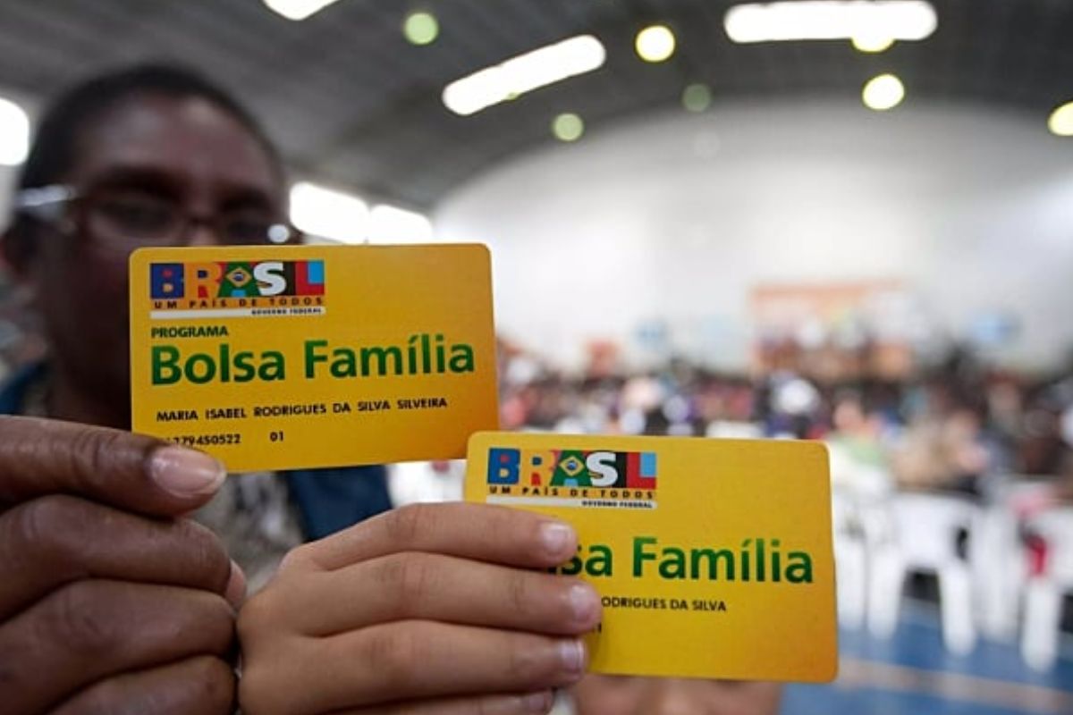 Um pai e seu filho seguram o cartão amarelo do benefício, eles estão felizes, pois o Bolsa Família amplia valores com benefícios para infância