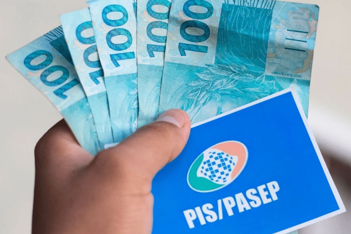 Em uma mão está o cartão do PIS/PASEP e a mesma mão segura algumas notas de R$ 100 após o pagamento da antecipação do PIS/Pasep 2025